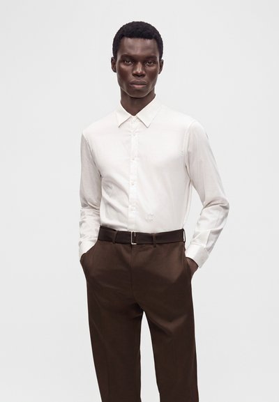 Homme portant une chemise blanche à manches longues boutonnée rentrée dans un pantalon marron foncé, debout les mains dans les poches devant un fond uni.