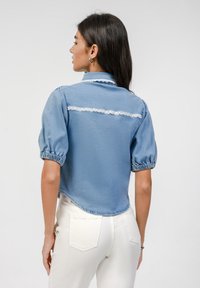 Blue denim shirt with puff sleeves, lace accent on the back, cropped design, and textured fabric. Paired with white jeans.
