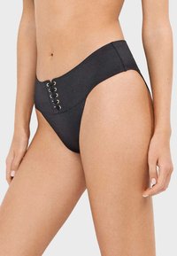 Femme portant un bas de bikini noir taille haute avec un détail de laçage œillet doré à l’avant, montrant le ventre et le haut des cuisses.