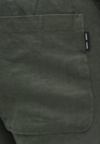 Tecido de linho verde com um bolso que apresenta uma etiqueta com o nome "Samsøe Samsøe". A textura é ligeiramente rugosa, com detalhes de costura visíveis.