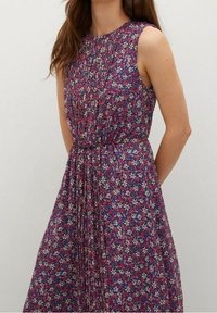 Robe florale violette au design sans manches, dotée de fins plis et d'une taille froncée. Le matériau a une finition douce et texturée avec de petites fleurs blanches.