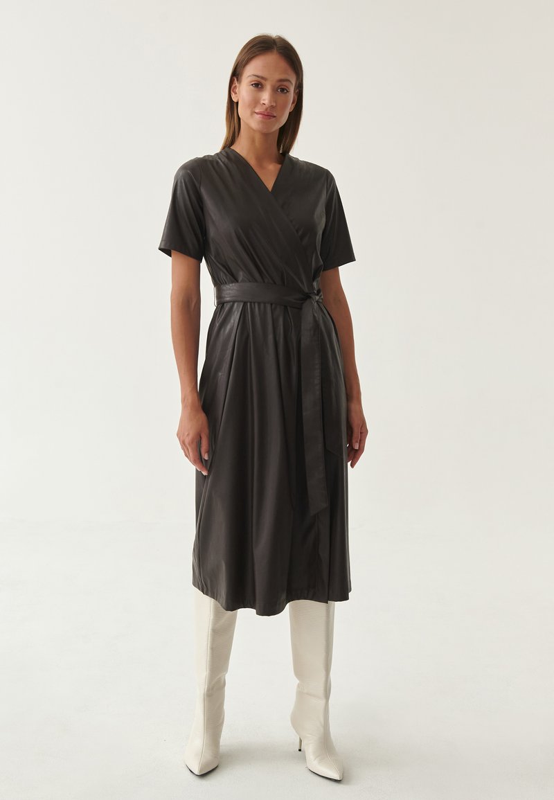 Abito nero di stile avvolgente in un materiale liscio e lucente, con maniche corte e vita regolabile, abbinato a stivali bianchi al ginocchio.