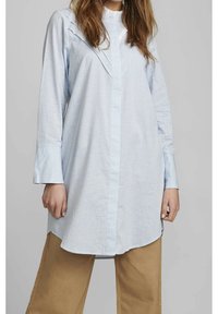 Ljusblå skjorta med knappar och rutmönster, veckdetaljer på axlarna, långa ärmar och rundad nederkant. Matchad med beige byxor.