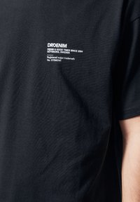 Camiseta de algodón negra con un pequeño logo blanco impreso en el pecho. La tela tiene una textura suave y un diseño clásico de cuello redondo.