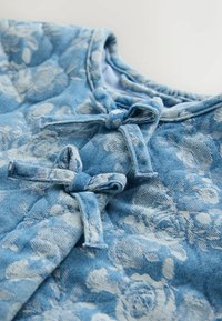 Džínový top světle modré barvy s květinovým vzorem. Má tři látkové mašle u límce a měkký texturovaný povrch s kulatým límečkem.