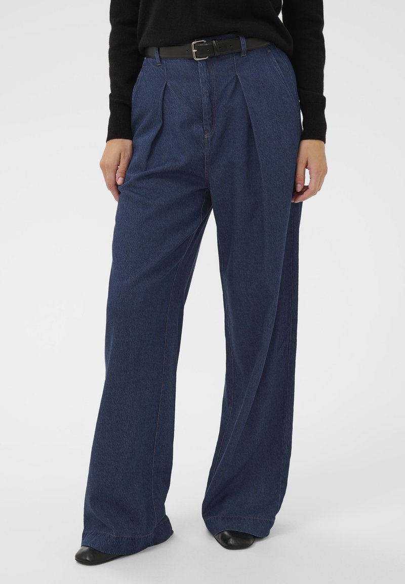 Széles szárú sötétkék farmernadrág, melynek derékrészén ráncok találhatók, fekete övvel és fekete cipővel viselve, fekete hosszú ujjú felsővel kombinálva.