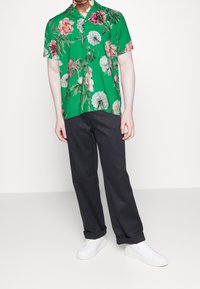 Camicia verde a maniche corte con un motivo floreale in rosa e bianco, abbinata a pantaloni scuri e scarpe da ginnastica bianche.