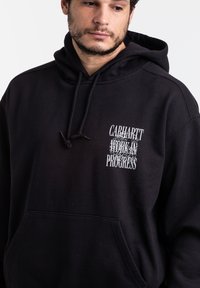 Felpa nera in cotone con tasca frontale, cappuccio con cordino e grafica testuale bianca recante la scritta "CARHARTT WORK IN PROGRESS" in font vari.