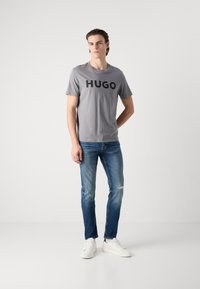 Tricou gri cu text mare negru "HUGO", combinat cu blugi albaștri. Modelul stă pe un fundal simplu, purtând adidași albi.