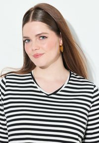 Černobíle pruhovaná halenka s výstřihem do V, vyrobená z měkké látky. Modelka má na sobě žluté náušnice ve tvaru listů a dlouhé, rovné vlasy.