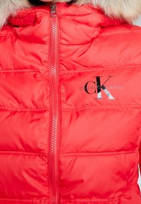 Rode pufferjas met een ritssluiting aan de voorkant, voorzien van een met bont gevoerde capuchon en het Calvin Klein Jeans-logo. Gequilte textuur en gladde stof.