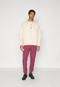 Smėlio spalvos džemperis su apykakle, išsiuvinėtu logotipu ir elastingu apačios kraštu. Derinamas su bordo spalvos sportinėmis kelnėmis su šoninėmis juostomis ir dėmėmis. Juodi batai.