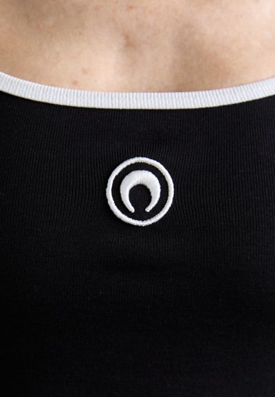 Črn rebrast material z belo ovratnikom in okroglo izvezeno logotipko v beli in črni barvi na sredini.