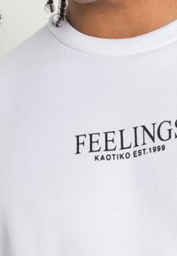 T-shirt bianca in cotone con colletto a costine, con la scritta "FEELINGS KAOTIKO EST. 1999" stampata in grassetto nero sulla parte frontale.