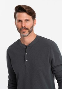 Uomo con capelli corti castani e barba curata, che indossa una camicia a maniche lunghe grigia scura con bottoni, guarda leggermente a destra con un'espressione neutra.