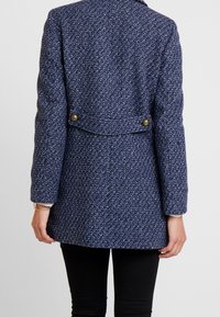 Kvinna iklädd en strukturert marinblå herringbone-kappa med två mässingsknappar på bakfältet, i kombination med svarta byxor.