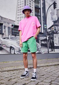 Młoda osoba w różowej koszuli, zielonych szortach, fioletowej czapce i w kratkowanych trampkach stoi na kostkowanej ulicy otoczona bańkami mydlanymi.