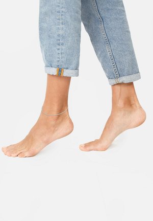 Braccialetto alla caviglia su un piede nudo, accanto a jeans di denim blu chiaro arrotolati con dettagli di cucitura arancione sui polsini. Il piede appare in una posizione rilassata.