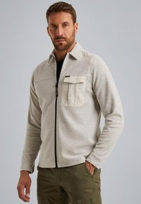 Lichtgrijze zip-up jack van een textuurstof, met een enkele borstzak in crème en een zwarte rits. Gecombineerd met olijfgroene broek.