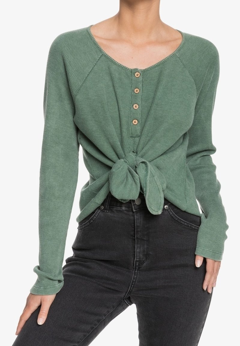 Femme portant un haut côtelé vert à manches longues avec quatre boutons et un lien devant, associé à un jean noir taille haute.