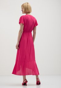 Femme aux cheveux blonds courts portant une robe midi rose vif et des sandales à talons rouges, se tenant devant un fond clair uniforme.