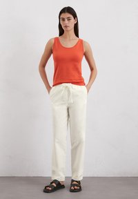 Oranges Tanktop mit Rundhalsausschnitt, kombiniert mit lockeren, cremigen Hosen mit Kordelzugbund, ergänzt durch schwarze Sandalen mit goldenen Akzenten.