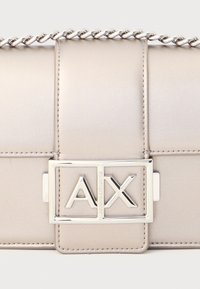 Metallisk beige handväska med kedjesnöre, med en rektangulär silverlås med "AX"-logotyp. Slät textur och sömdetaljer synliga.