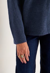 Marineblauer Strickpullover mit gerippten Bündchen, kombiniert mit dunkler Jeans. Die Hand ziert drei goldene Ringe mit glatten und strukturierten Designs.
