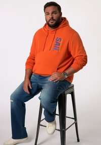 Felpa arancione con testo blu "SAIL", occhielli metallici e cordini. Indossata con jeans chiari strappati e sneakers bianche, seduto su uno sgabello.