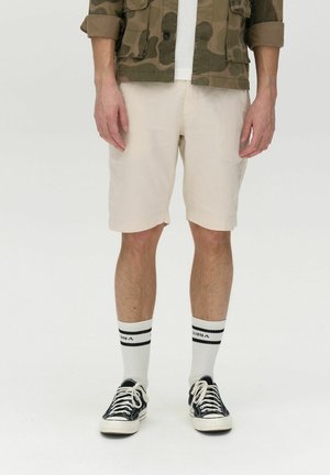 Shorts bej de lungime la genunchi dintr-un material ușor, asortați cu șosete cu dungi alb-negru și pantofi din pânză negri. Modelul poartă o jachetă de camuflaj.