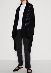 Svart öppen cardigan med strukturerade paneler, i kombination med en vit skjorta och mörka jeans. Har en avslappnad silhuett och är tillverkad av ett lätt stickat material.