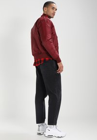 Giacca bomber rossa con una texture liscia, abbinata a una camicia a quadri sotto e jeans neri larghi, completata da sneakers bianche.