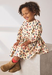 Floral-patterned, cream corduroy dress with long sleeves and gathered waist. Brown ankle boots with a textured finish and red socks are worn.