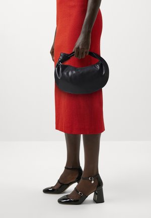 Personne en robe rouge tenant un sac à main en cuir noir, portant des chaussures Mary Jane vernies noires à talons carrés.