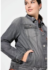 Harmaa farkkutakki, jossa on kaulus, etupainonappi kiinnitys, kaksi rintataskua ja hienovarainen kulunut yksityiskohta. Pitkät hihat ja sivutaskut.