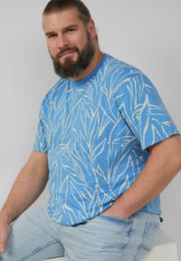 Blaues T-Shirt mit kurzen Ärmeln und weißem Blattmuster, mit Rundhalsausschnitt. Getragen mit hellblauen Jeans.