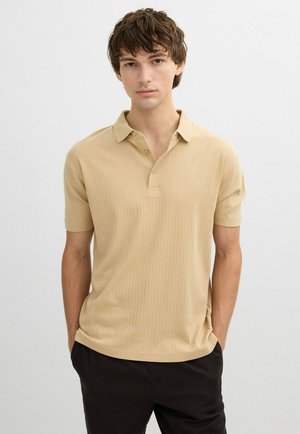 Joven con una camisa tipo polo beige de manga corta con botones, de pie con las manos en los bolsillos de los pantalones negros contra un fondo blanco.