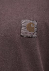 Tamno ljubičasta pamučna košulja s ušivenim zakrpom s kružnim logom i tekstom u kontrastnoj zlatnoj i tamnocrvenoj boji. Glatka tekstura, ležeran dizajn.