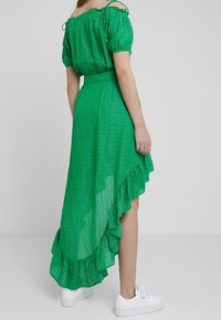 Vihreä ruudullinen mekko, jossa on olkapäiden päältä avautuva muotoilu, röyhelöhelma ja läpikuultava rakenne, yhdistettynä valkoisiin tennareihin.