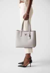 Sac en cuir gris avec deux poignées, accents métalliques et un petit détail de porte-clés, associé à un haut beige et un pantalon clair.