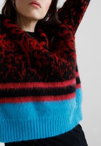 Sweter dzianinowy z wyrazistym czerwono-czarnym wzorem i niebieskim ściągaczem. Teksturowana tkanina dodaje głębi wzorowi.