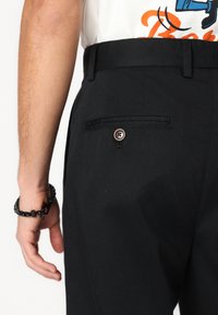 Calças pretas com uma textura suave, apresentando um bolso lateral com fecho de botão e passadores para cinto, combinadas com uma camisa branca.