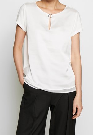 Vrouw die een witte blouse met korte mouwen draagt met een keyhole-halslijn en een cirkelvormige metalen ring, gecombineerd met zwarte, wijd uitlopende broek.