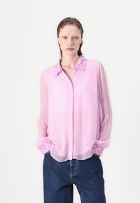 Rosane, durchsichtige Bluse mit weicher Textur. Verfügt über einen Rüschenausschnitt und elastische Ärmelbündchen. Kombiniert mit dunkelblauen Jeans.