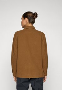 Bruine, langemouwen shirt met een kraag, gemaakt van textuurstof en met knoopmanchetten, gezien vanaf de achterkant tegen een lichte achtergrond.
