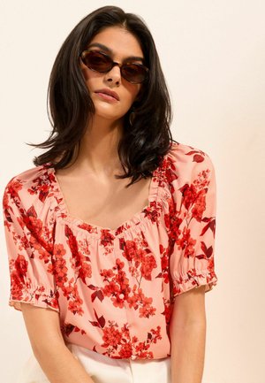 Žena s tmavými vlasmi po ramená, ktorá má na sebe oválne slnečné okuliare a ružovú blúzku s červeným kvetinovým vzorom na jednoliatom pozadí.