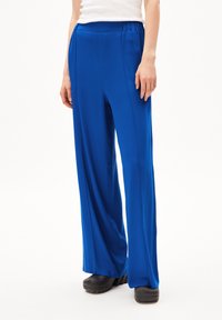 Blauwe wijde pantalon van gladde stof, met een elastische tailleband, verticale voornaad en een ontspannen pasvorm. G gedragen met zwarte schoeisel.