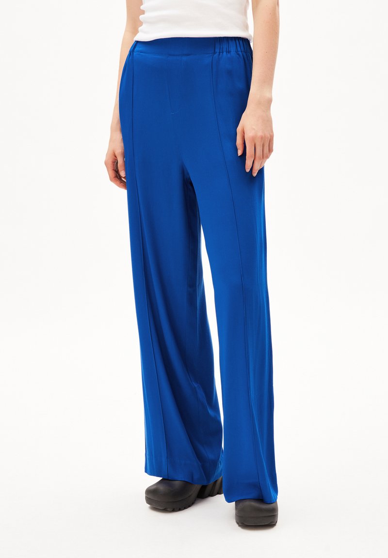 Blauwe wijde pantalon van gladde stof, met een elastische tailleband, verticale voornaad en een ontspannen pasvorm. G gedragen met zwarte schoeisel.