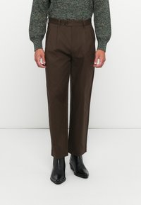 Man wearing brown straight-leg trousers, black pointed-toe boots, and a green knit sweater standing against a white background.