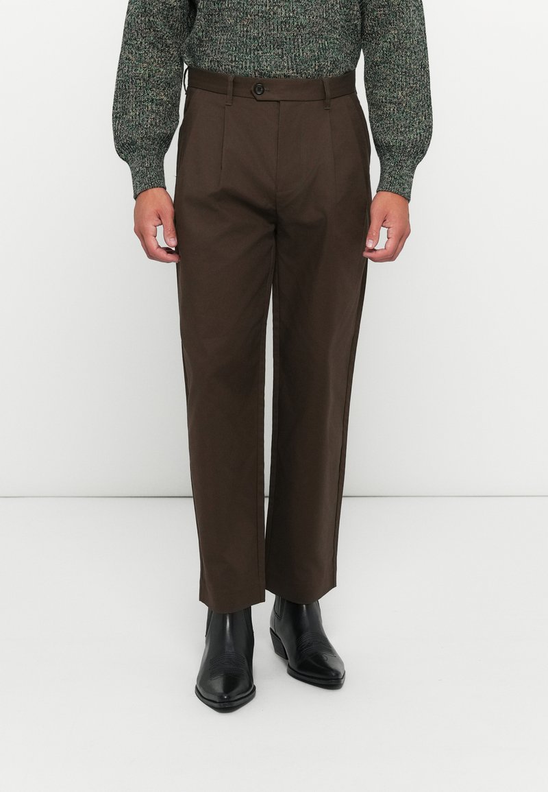 Man wearing brown straight-leg trousers, black pointed-toe boots, and a green knit sweater standing against a white background.